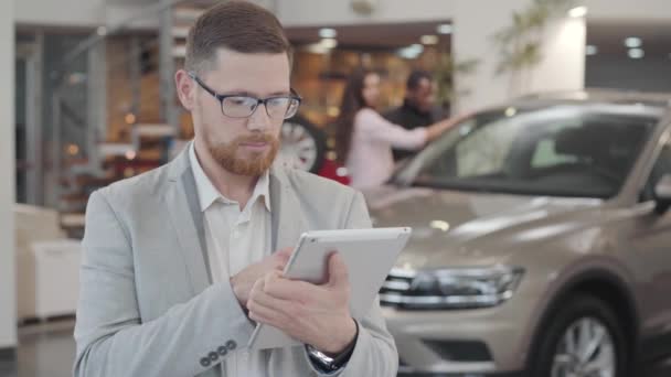 Retrato de cerca de un hombre caucásico serio revisando su tableta, mirando a la cámara y haciendo un gesto afirmativo agitando la cabeza. Confiado concesionario de coches de éxito en el fondo del nuevo automóvil en la sala de exposición . — Vídeos de Stock