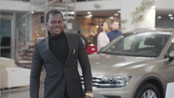 Kendine güvenen Afrikalı Amerikalı adam kameraya geliyor, yeni otomobilin anahtarlarını gösteriyor ve evet diyor. Araba galerisinde siyah takım elbiseli tatmin olmuş bir iş adamı. Araba işi.. — Stok video