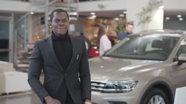 Empresário afro-americano positivo mostrando as chaves do carro na câmera e gesticulando sim. Jovem homem feliz em terno preto comprando automóvel no showroom. Concessionária de automóveis, empresa de automóveis . — Vídeo de Stock