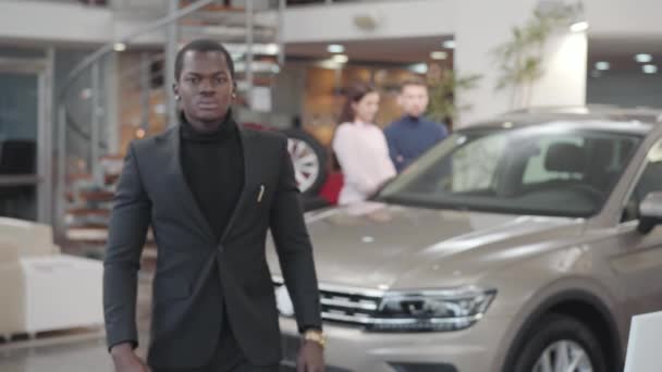 Porträtt av stilig afroamerikansk bilhandlare som visar bilnycklar framför kameran och gester ja. Ung glad säljare i svart kostym säljer bil i showroom. Bilhandel, bilindustri. — Stockvideo