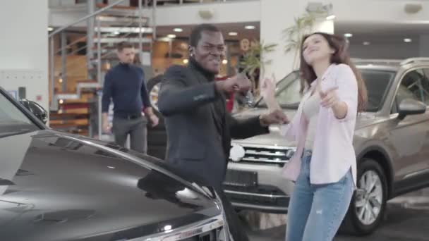 Fröhlicher afrikanisch-amerikanischer Mann und schöne kaukasische Frau tanzen neben dem neuen Auto mit Autoschlüssel. zufriedenes multiethnisches Paar prahlt mit seinem Kauf. Autohaus, Autohaus. — Stockvideo