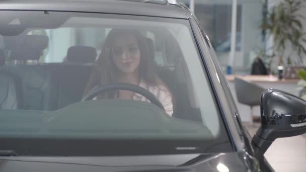Portret van een jonge, mooie blanke vrouw in de nieuwe auto in de showroom. Zelfverzekerd lachend meisje kiezen auto te kopen. Autohandel, autohandel. — Stockvideo