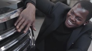 Yeni siyah otomobilin tamponunu ve araba kaputunu okşayan mutlu Afrikalı Amerikalı adamın portresi. Başarılı genç işadamı sergi salonundaki alımına hayran. Araba bayiliği, araba işi..