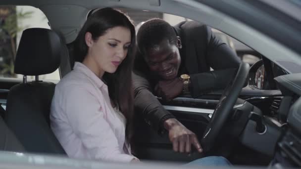 Young Caucasian woman sitting in car salon and talking with African American car dealer. Beautiful successful businesslady choosing automobile in showroom. Car dealership, car business. — ストック動画