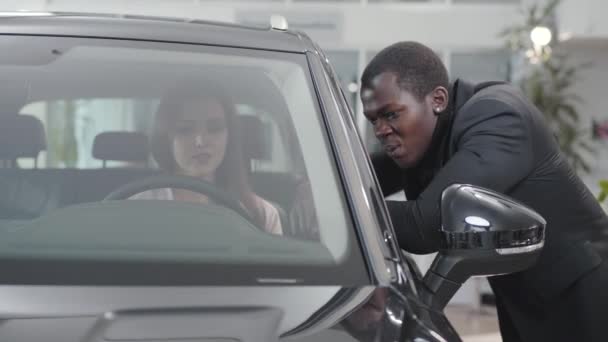 Porträtt av leende afroamerikansk bilhandlare som pratar med en ung vit kvinna i bilsalong. Professionell hjälpa kunden att välja bästa bil i showroom. Bilhandel, bilindustri. — Stockvideo