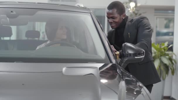 Confiado comerciante masculino afroamericano hablando con la joven mujer caucásica sentada en el salón de automóviles. Vendedor exitoso ayudando al cliente a elegir el automóvil en la sala de exposición. concesionario de coches, negocio de automóviles . — Vídeos de Stock