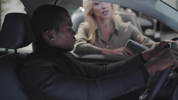 Блондинка-продавщица автомобилей белого цвета, объясняющая характеристики афроамериканскому покупателю, сидящему в автосалоне. Успешный молодой человек выбирает автомобиль в выставочном зале. Автосалон, автобизнес . — стоковое видео