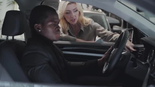 成功的非裔美国人坐在汽车沙龙，与白人女性汽车经销商交谈。 年轻自信的家伙选择在展示厅的汽车。 汽车经销商，汽车生意. — 图库视频影像