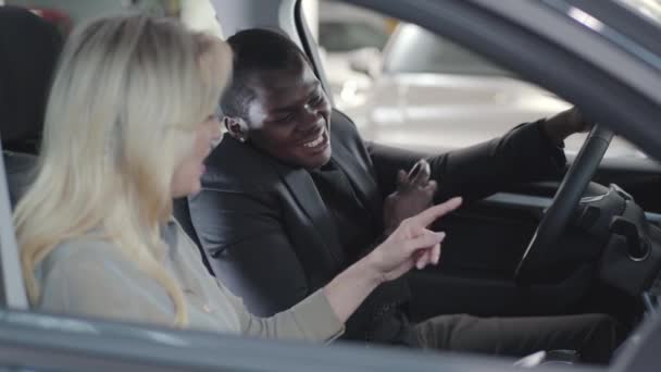 Vista lateral do homem afro-americano sorridente segurando as chaves do carro e ouvindo o revendedor de carros caucasiano. Vendedora loira e cliente sentado no salão de beleza do carro. Comerciante que vende automóveis. Negócios de automóveis . — Vídeo de Stock