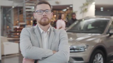 Kafkasyalı başarılı bir araba satıcısının portresi kollarını kavuşturup başını sallayarak evet işareti yapıyor. Satıcı kameraya bakıyor ve gülümsüyor. Arka planda yeni arabalarına hayran olan çok ırklı bir çift var..