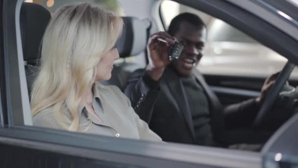 아름다운 금발의 백인 여성에게 자동차 열쇠를 주는 아프리카 계 미국인 상인의 흐릿 한 모습. 성공 한 사업가인 그녀는 카메라를 보고 구매를 자랑 했습니다. 자동차 대리점, 자동차 사업. — 비디오