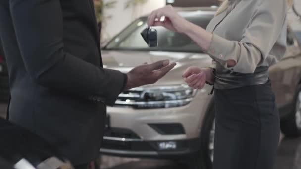 Primer plano de dos personas estrechando la mano en el fondo de los automóviles nuevos. Mano afroamericana masculina que toma las llaves del coche de la vendedora caucásica. concesionario de coches, negocio de automóviles . — Vídeos de Stock