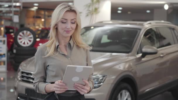 Confiante jeune femme caucasienne utilisant une tablette, regardant la caméra et souriant. Succès concessionnaire de voitures blondes posant à l'arrière-plan de nouvelles voitures. Concessionnaire automobile, entreprise automobile . — Video