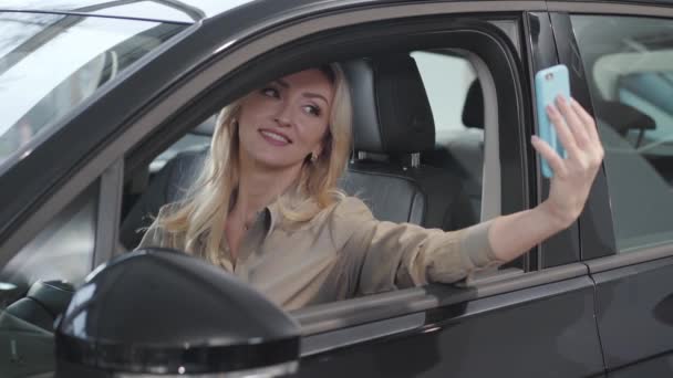 Mulher branca loira bonita a tirar selfie no salão do carro novo. Empresária bem sucedida tirando fotos no automóvel comprado no showroom. Concessionária de automóveis, empresa de automóveis . — Vídeo de Stock