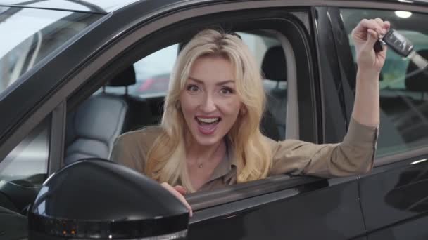 Mujer caucásica feliz con el pelo rubio sentado en el salón y presumiendo de las llaves del coche. Exitosa empresaria comprando automóviles en la sala de exposición. concesionario de coches, negocio de automóviles . — Vídeo de stock