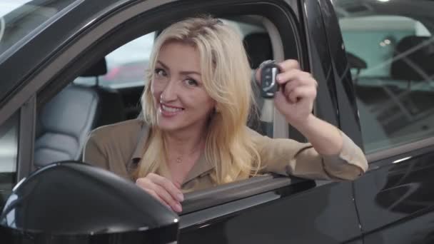 Mujer rubia caucásica satisfecha con ojos marrones alardeando de las llaves del coche mientras se sienta en el salón. Exitosa empresaria comprando automóviles en la sala de exposición. concesionario de coches, negocio de automóviles . — Vídeos de Stock