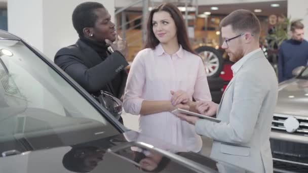 Αυτοπεποίθηση Καυκάσιος έμπορος αυτοκινήτων στέκεται με τους πελάτες δίπλα στο αυτοκίνητο και δείχνει τις προδιαγραφές στο tablet του. Πολυφυλετικό ζευγάρι αγοράζει νέο αυτοκίνητο στην έκθεση. αντιπροσωπεία αυτοκινήτων, επιχείρηση αυτοκινήτων. — Αρχείο Βίντεο