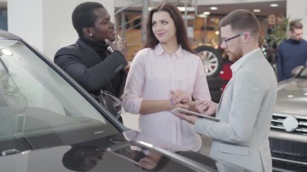 Confident Caucasian car dealer standing with clients next to the automobile and showing specifications on his tablet. Multiracial couple buying new car in showroom. Car dealership, car business. — ストック動画