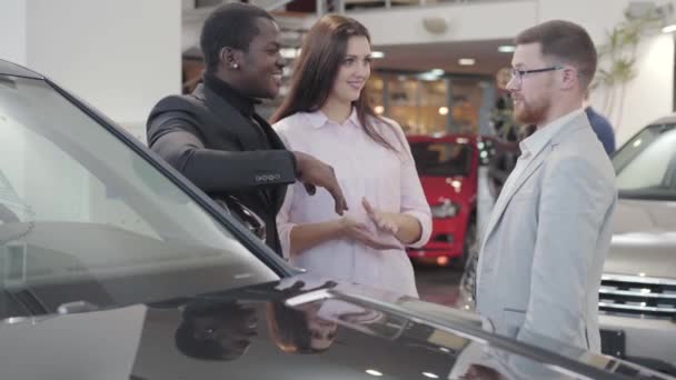 Young Caucasian salesman giving car keys to biracial couple in showroom. Beautiful young woman hugging positive African American man, positive guy starting dancing. Car dealership, car business. — ストック動画