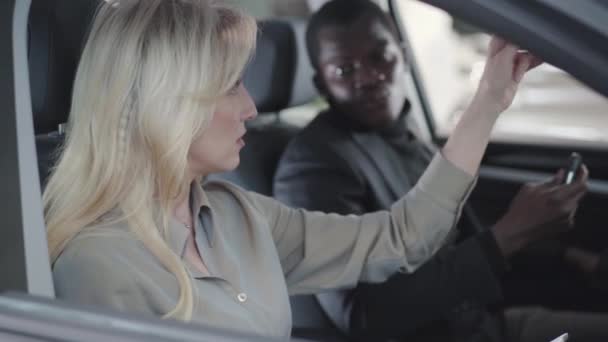 侧视图美丽的金发白人妇女坐在汽车沙龙与平板，并向非洲裔美国客户解释规格。 汽车经销商出售汽车。 汽车经销商，汽车生意. — 图库视频影像