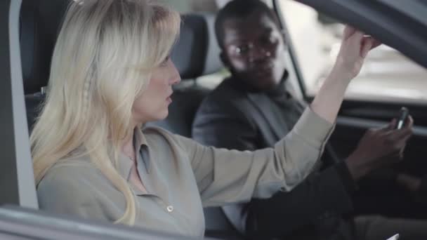 Vista lateral de la hermosa mujer rubia caucásica sentada en el salón de automóviles con tableta y explicando las especificaciones al cliente afroamericano. Distribuidor de coches de venta de automóviles. concesionario de coches, negocio de automóviles . — Vídeo de stock