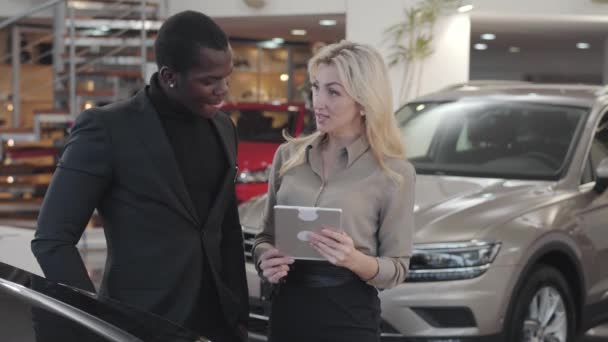 Femme blanche adulte aux cheveux blonds tenant la tablette et expliquant les spécifications à l'homme afro-américain. Femme concessionnaire de voiture debout avec le client à côté de l'automobile. Concessionnaire automobile, entreprise automobile . — Video