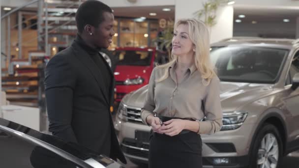 Mooie blanke vrouw geeft autosleutels aan de Afro-Amerikaanse man in de showroom. Gelukkige cliënt gebaart ja en glimlachende verkoopster klapt in de handen. Autohandel, autohandel. — Stockvideo