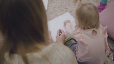Kafkasyalı genç bir kadın ve renkli kalemlerle çizim yapan küçük bir kız bebek manzarası. Anne, sevimli kızını oynuyor ve eğitiyor. Hamileliğin neşesi. Omzundan vuruluyor..