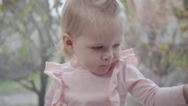 Närbild porträtt av söt vit liten flicka med grå ögon och blont hår sitter på fönsterbrädan. Barn i rosa blus sträcker ut händerna till kameran och äter äpple. Lycklig barndom. — Stockvideo