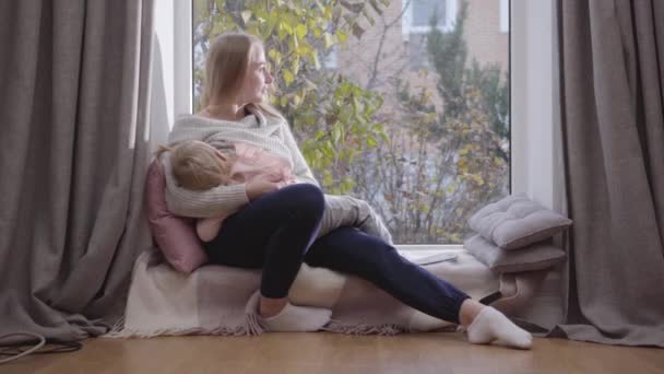 Portrait of young happy Caucasian woman sitting on low windowsill and holding her daughter on hands. Smiling mother looking at her child and talking. Family spending autumn day together at home. — Stock Video