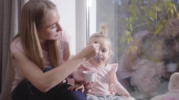 Porträtt av en vit ung kvinna som sitter på en låg fönsterbräda och matar en liten blond flicka. Vacker mor tar hand om sin bedårande dotter. Glädje i moderskapet, lycklig barndom. — Stockvideo