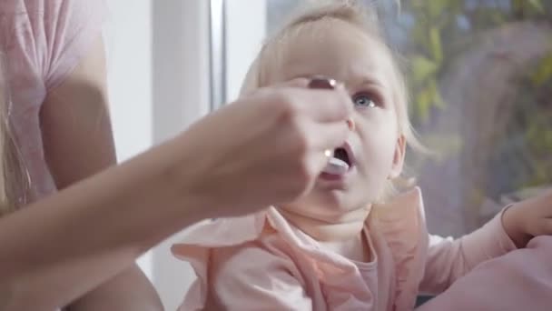 Faccia da primo piano di una ragazzina caucasica che mangia cibo per bambini. Madre che nutre il suo bambino affascinante con occhi grigi profondi. Felice maternità, gioia della maternità . — Video Stock