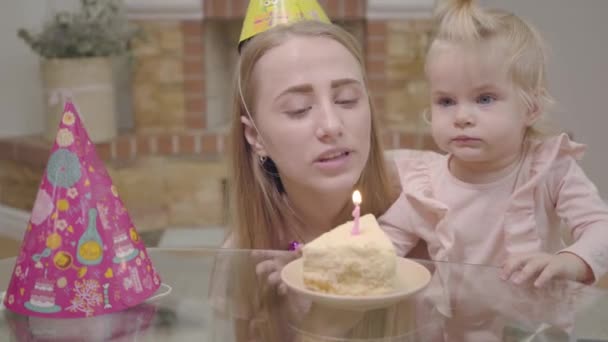 Primer plano de la joven mujer caucásica en sombrero de fiesta y niña pequeña soplando vela en el pedazo de pastel. Encantadora niña celebrando su primer cumpleaños con su madre. Feliz infancia, alegría de la maternidad . — Vídeo de stock