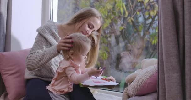 Portret van klein blond kind zittend op moeders benen en tekenen met kleurrijke potloden. Jonge glimlachende vrouw die haar dochtertje bewondert en haar hoofd strelt. Bioscoop 4k beelden Prores Hq. — Stockvideo