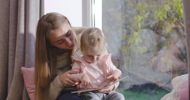 Close-up van Kaukasische moeder en baby meisje zitten bij vensterbank en lezen boek. Jonge aantrekkelijke vrouw die thuis een dochter opvoedt. Gelukkige kindertijd, vreugde van het moederschap. Bioscoop 4k beelden Prores Hq. — Stockvideo