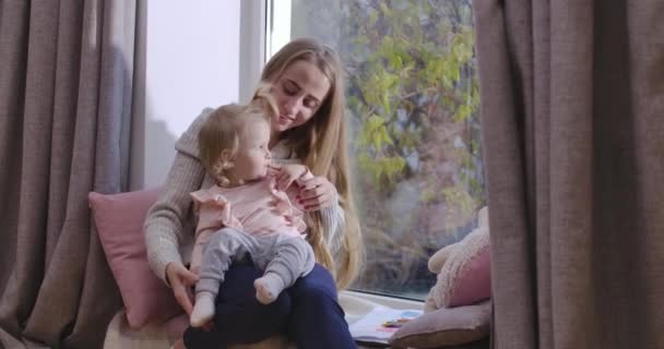 Junge schöne kaukasische Mutter und Tochter sitzen auf der Fensterbank und schauen aus dem Fenster. Frau spricht mit ihrem Kind und zeigt etwas im Freien. Kino 4k Filmmaterial prores hq. — Stockvideo