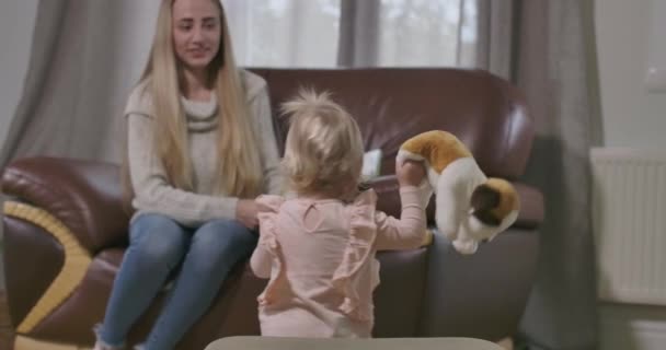 Retrato de criança caucasiana bonito tomando cachorro de brinquedo macio e indo para mulher sorridente sentado no fundo. Mãe feliz admirando sua filhinha andando sozinha. Cinema 4k footage ProRes HQ . — Vídeo de Stock