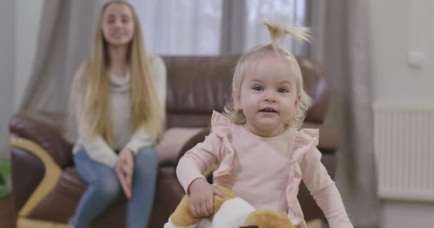 Közelkép egy kedves kaukázusi kislányról, aki kamerába néz és mosolyog. Csinos gyerek kezében játékkutya. Az elmosódott anyja ül a háttérben és csodálja a lányát. Mozi 4k felvételek Prores Hq. — Stock videók