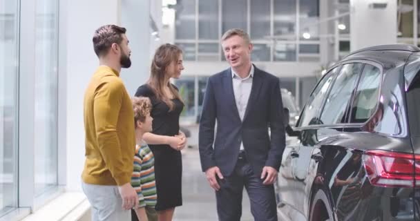 Homme caucasien adulte en costume d'affaires congradant jeune famille avec l'achat d'une nouvelle automobile dans le concessionnaire automobile. Garçon joyeux avec les cheveux bouclés acheter une voiture avec les parents. Cinéma séquences 4k ProRes — Video