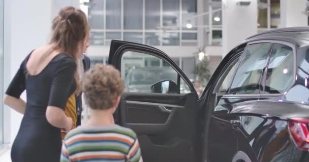 Mosolygó kaukázusi autókereskedő kinyitja az autó ajtaját az autószalonban. Fiatal szakállas vásárló keresi a szalont és beszél a feleségével és a fiával. Boldog család vesz új autót. Mozi 4k felvételek Prores Hq. — Stock videók