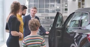 Genç, sakallı, beyaz bir adam kuaföre gidip oğullarına fikrini soruyor. Küçük çocuk annesinin yanında duruyor ve parmağını gösteriyor. Mutlu aile yeni araba alıyor. Araba işi. Sinema 4k görüntüleri Prores Hq.