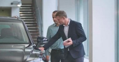 Kareli gömlekli müşteriye tabletle araba kapısı açan kendine güvenen beyaz erkek tüccar. Başarılı bir adam yeni otomobil seçiyor. Araba galerisi, araba işi. Sinema 4k görüntüleri Prores Hq.