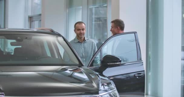 两名成熟的白人男子站在黑色汽车旁边握手。 男汽车经销商在汽车展示室与顾客做交易. 汽车经销商，汽车生意。 电影4k镜头Prores Hq. — 图库视频影像