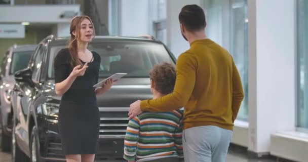 Ung säker kaukasisk kvinna som förklarar bilens specifikationer för manlig klient. Baksidan av vuxen man väljer ny bil med son. En framgångsrik kille som köper fordon. Film 4k Prores Hq. — Stockvideo