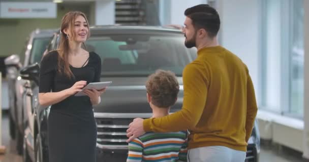 Baksidan bild av vuxna kaukasiska mannen väljer bil med son i bilhandlare. Kvinnlig bilhandlare som håller i surfplattan och pratar med kunder. Bilhandlare, bilfirma. Film 4k Prores Hq. — Stockvideo