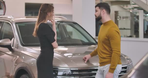 若い白人のカップルは、車の販売代理店に立って、新しい自動車を選択します。自動車ショールームで車を買う成功した男と女。車のディーラー、車のビジネス。映画4k映像｜Prores Hq. — ストック動画