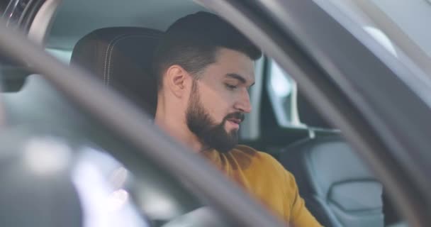 Retrato del joven hombre caucásico en suéter de mostaza sentado en el salón de coches y hablando. Cliente barbudo con éxito la elección de un nuevo automóvil para comprar en la sala de exposición. Negocios de autos. Cinema 4k metraje ProRes HQ . — Vídeo de stock