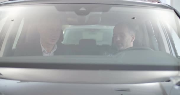 Twee volwassen blanke mannen zitten in een autosalon te praten. Man handelaar het maken van verkoop aankoop deal met volwassen klant. Schieten door de voorruit. Autohandel, autohandel. Cinema4k beelden Proreshq — Stockvideo