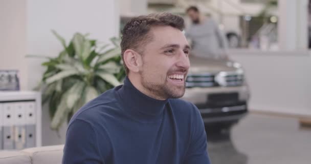 Κοντινό πρόσωπο ενός όμορφου Καυκάσου που κάθεται στην αντιπροσωπεία αυτοκινήτων και χαμογελάει. Νεαρός επιτυχημένος επιχειρηματίας που ξεκουράζεται μετά την επιλογή νέων αυτοκινήτων. Δουλειά αυτοκινήτου. Κινηματογράφος 4k πλάνα Prores Hq. — Αρχείο Βίντεο