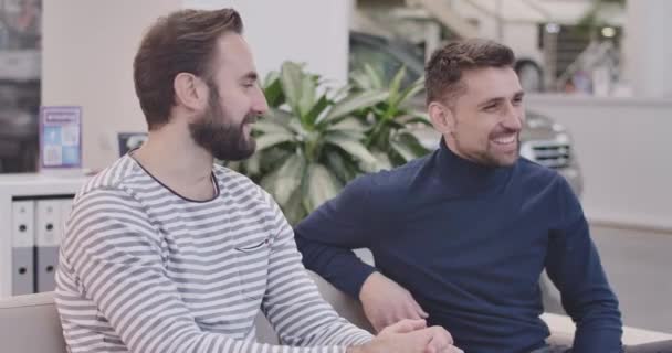 Vista lateral de dos hombres guapos caucásicos sentados en un concesionario de automóviles, sonriendo y hablando. Jóvenes empresarios discutiendo la compra de coches en la sala de exposición. Negocios de autos. Cinema 4k metraje ProRes HQ . — Vídeos de Stock